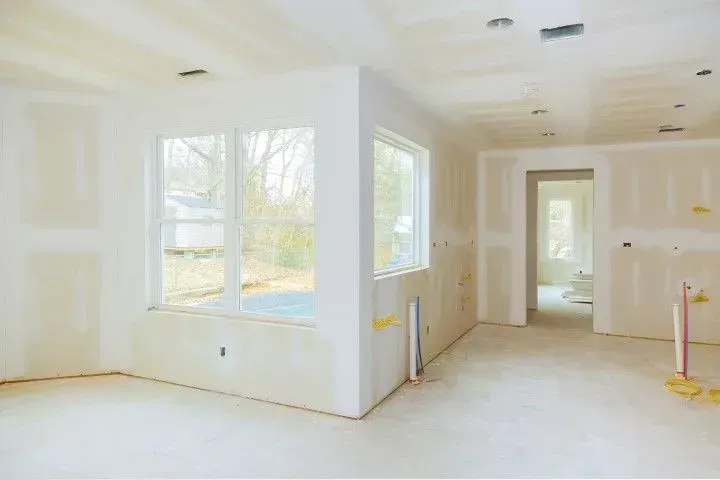 An interior view of a room under construction with unfinished drywall, exposed studs, and a large window.
