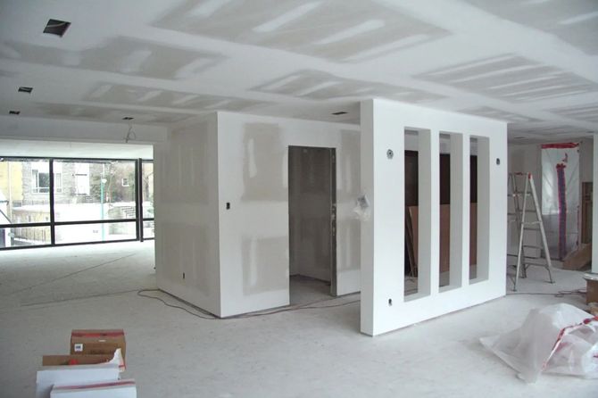 A construction worker on a ladder installs drywall on a wood-framed wall in an unfinished room.