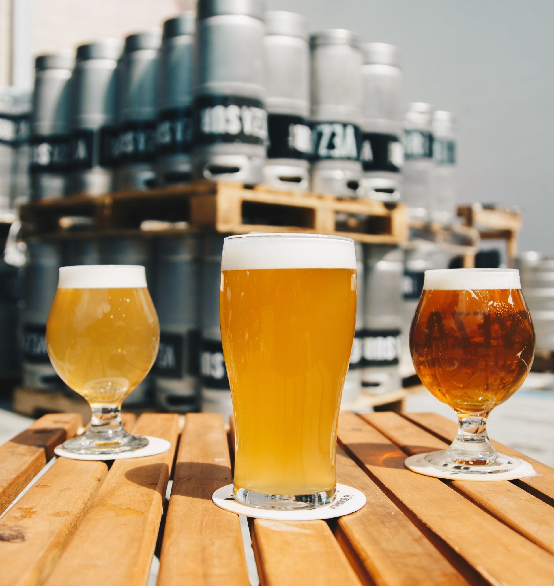 Three glasses of beer on a wooden table with kegs in the background
