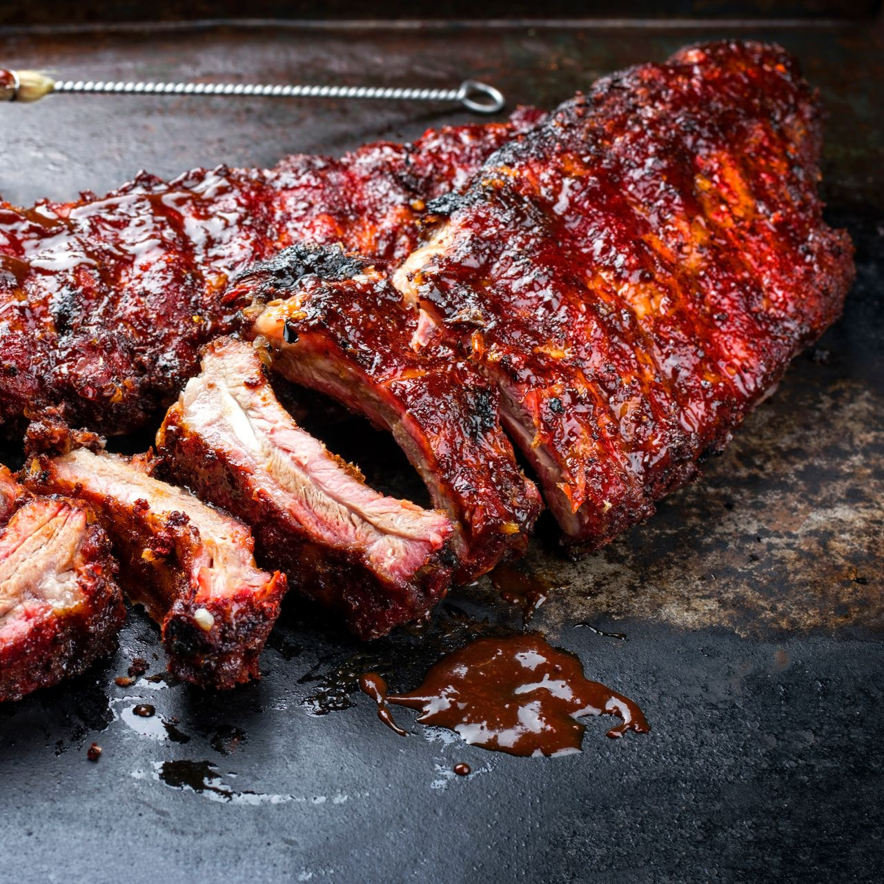 Rack of barbecued ribs on a dark metal surface, with a dark, glossy sauce.