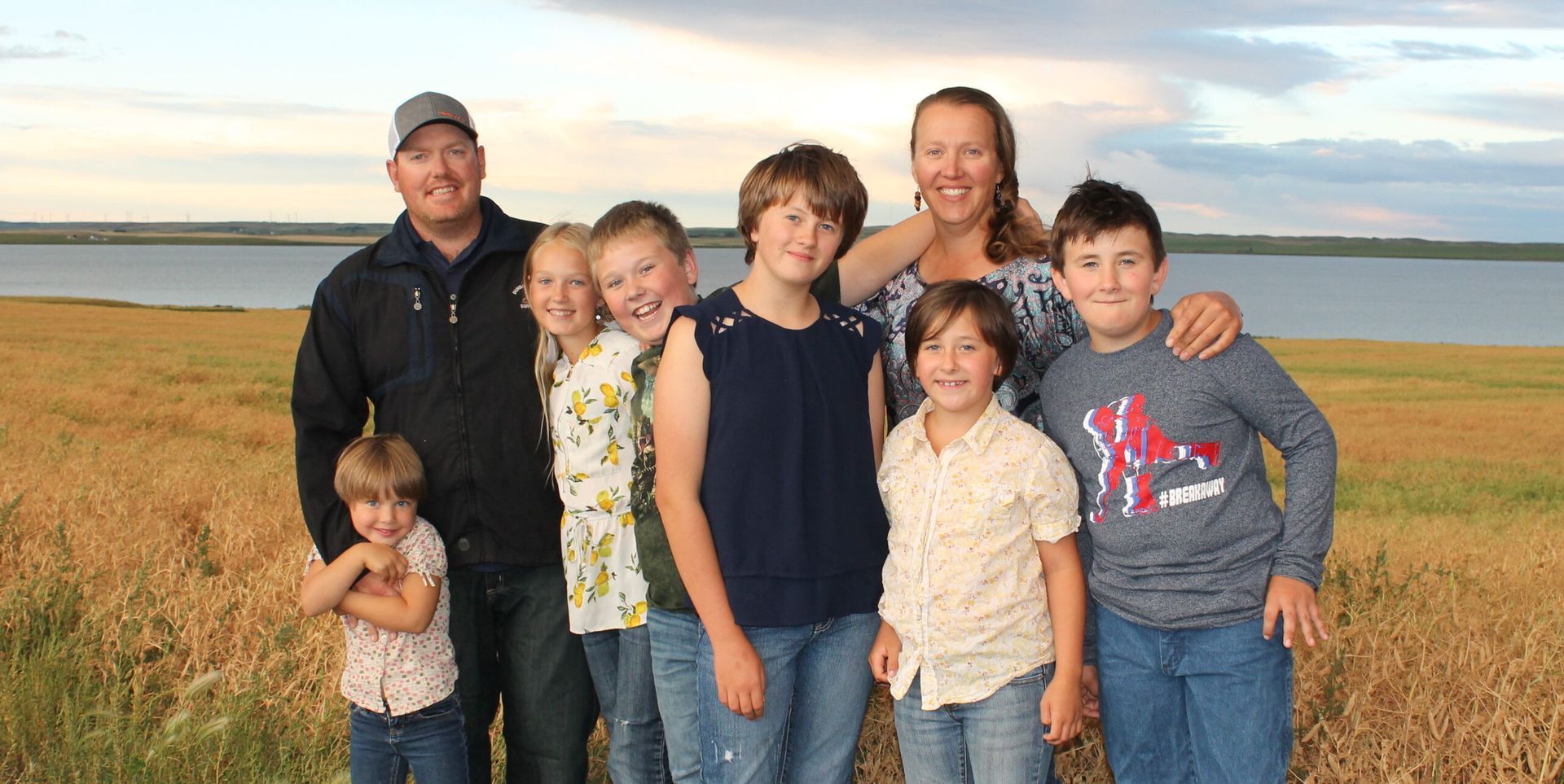 Family posing in a field of wheat near a body of water under a cloudy sky.