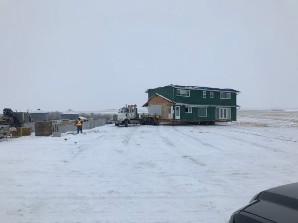 Two-story green house being moved by a truck on a snowy, open field. A person in orange vest stands near.