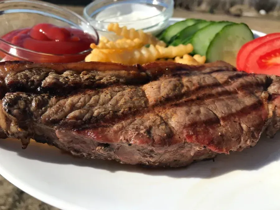 Grilled steak with grill marks, served with fries, cucumbers, tomato, ketchup, and white sauce.