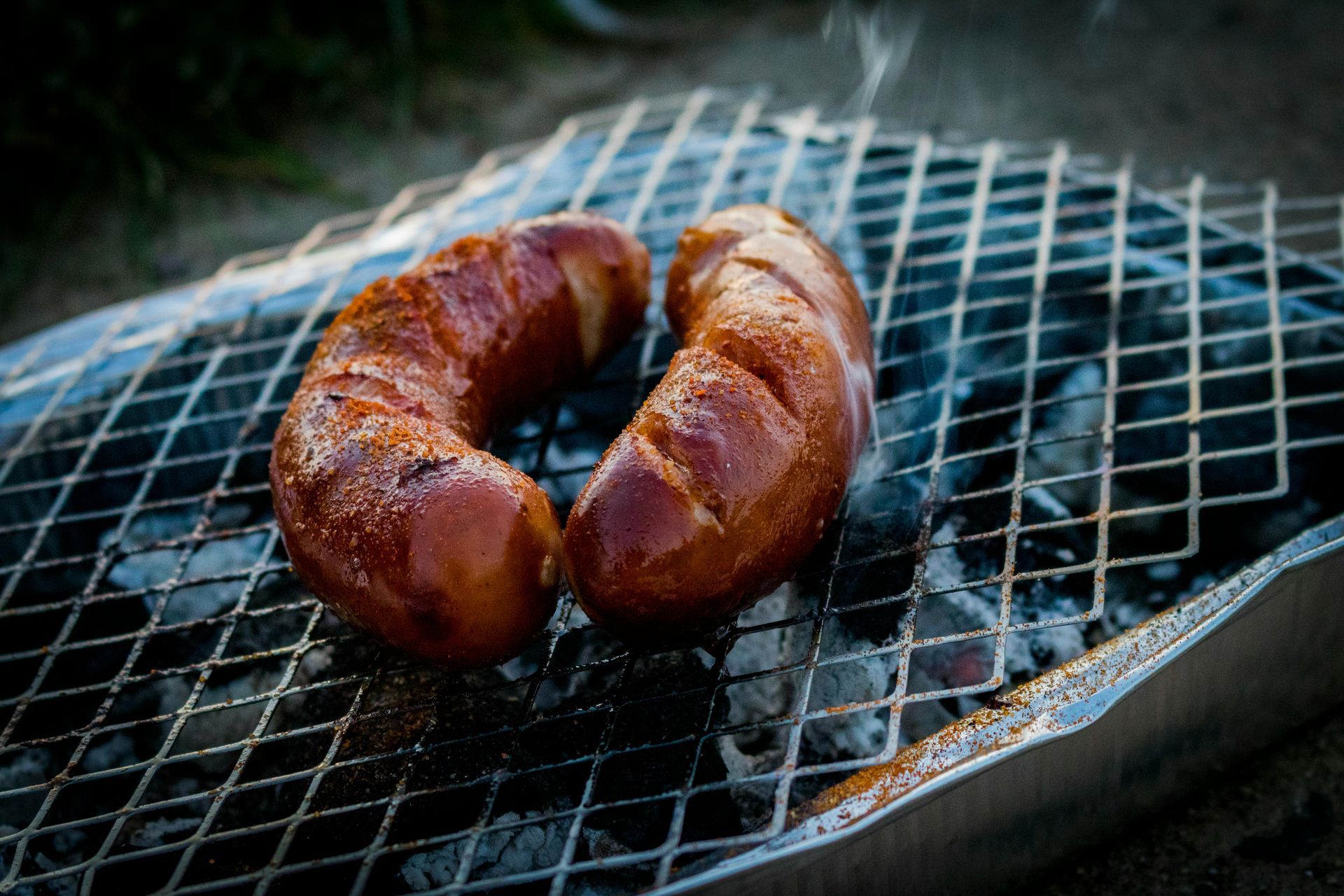 Two browned sausages grilling on a charcoal grill, with light smoke rising.