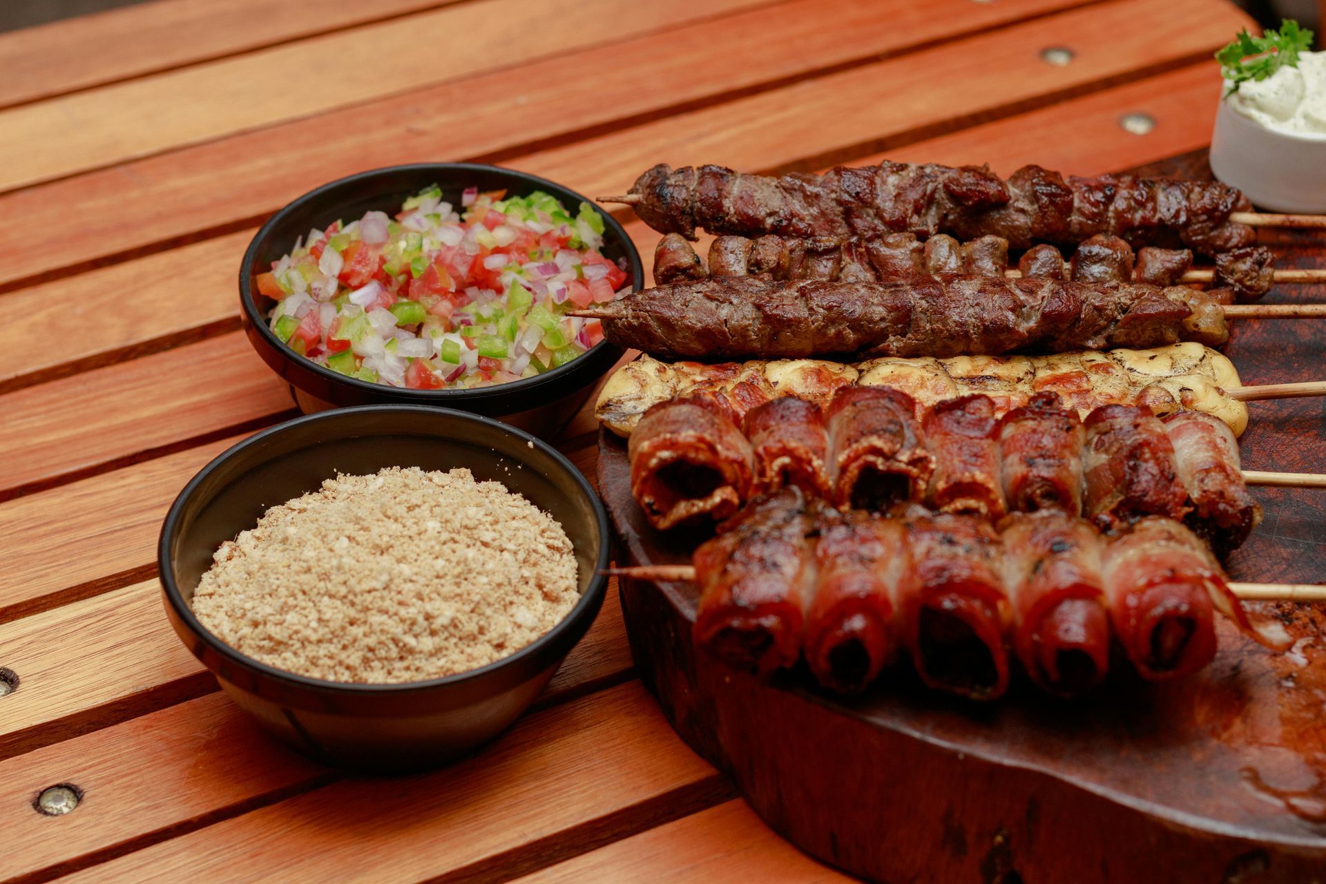 Grilled meat skewers, side dishes: salsa, seasoned flour, and cream, served on a wood surface.