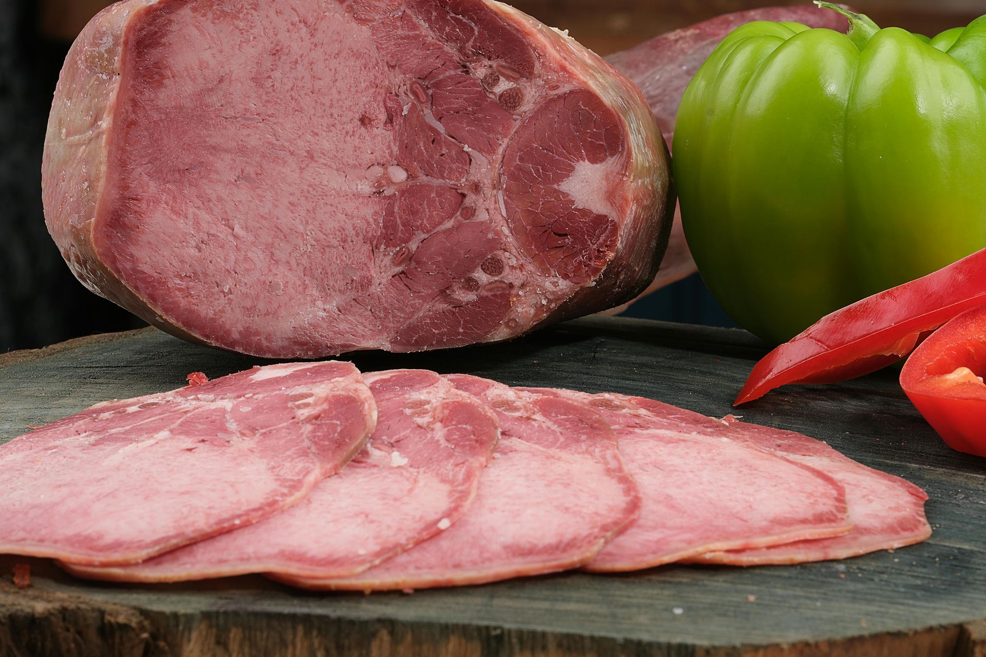 Sliced ham with a whole ham, green pepper, and red pepper on a wooden surface.