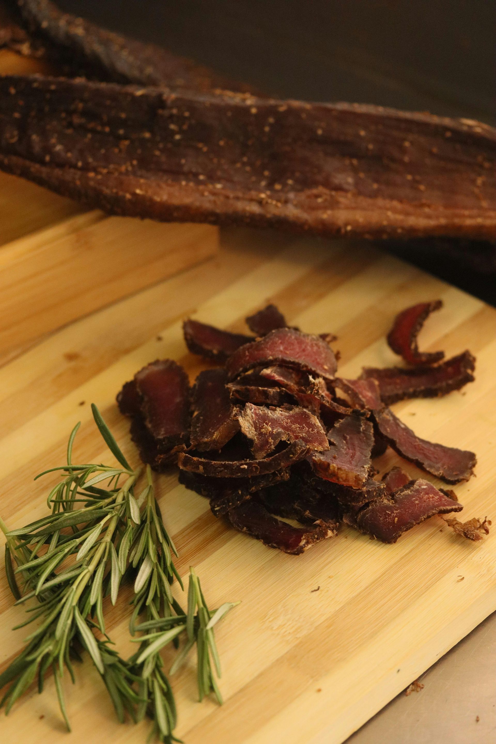 Sliced biltong with rosemary sprigs on a wooden cutting board; dried biltong pieces in the background.