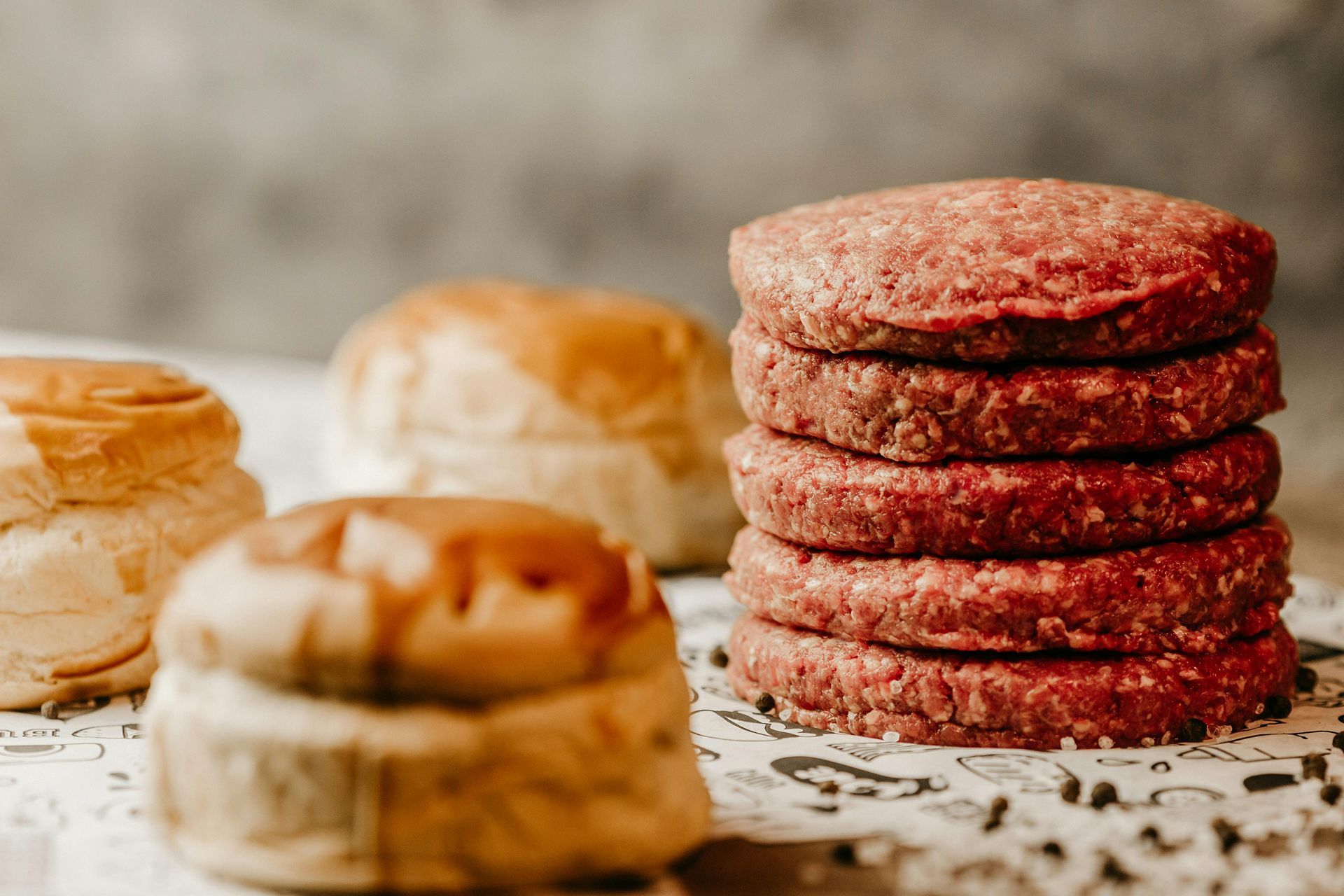 Raw hamburger patties stacked next to partially visible hamburger buns on a speckled surface.