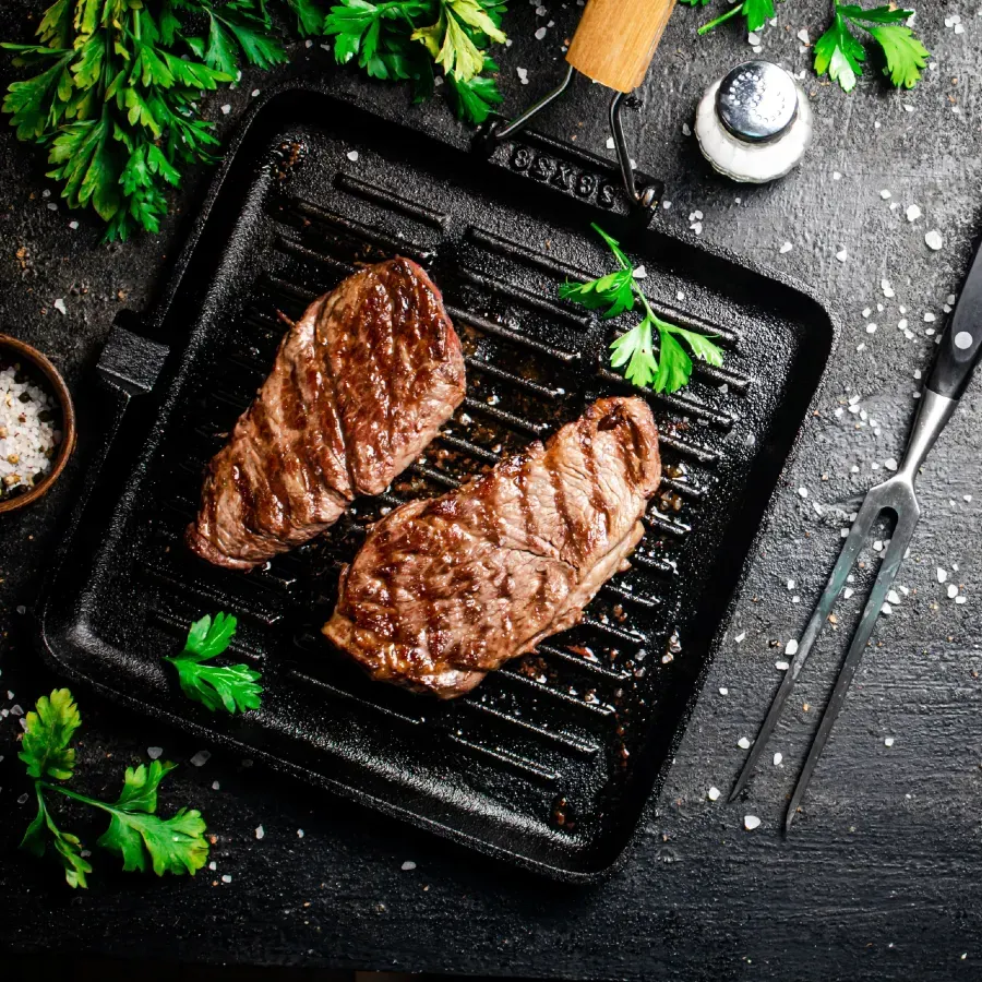 Two grilled steaks on a black griddle pan, garnished with parsley and salt.