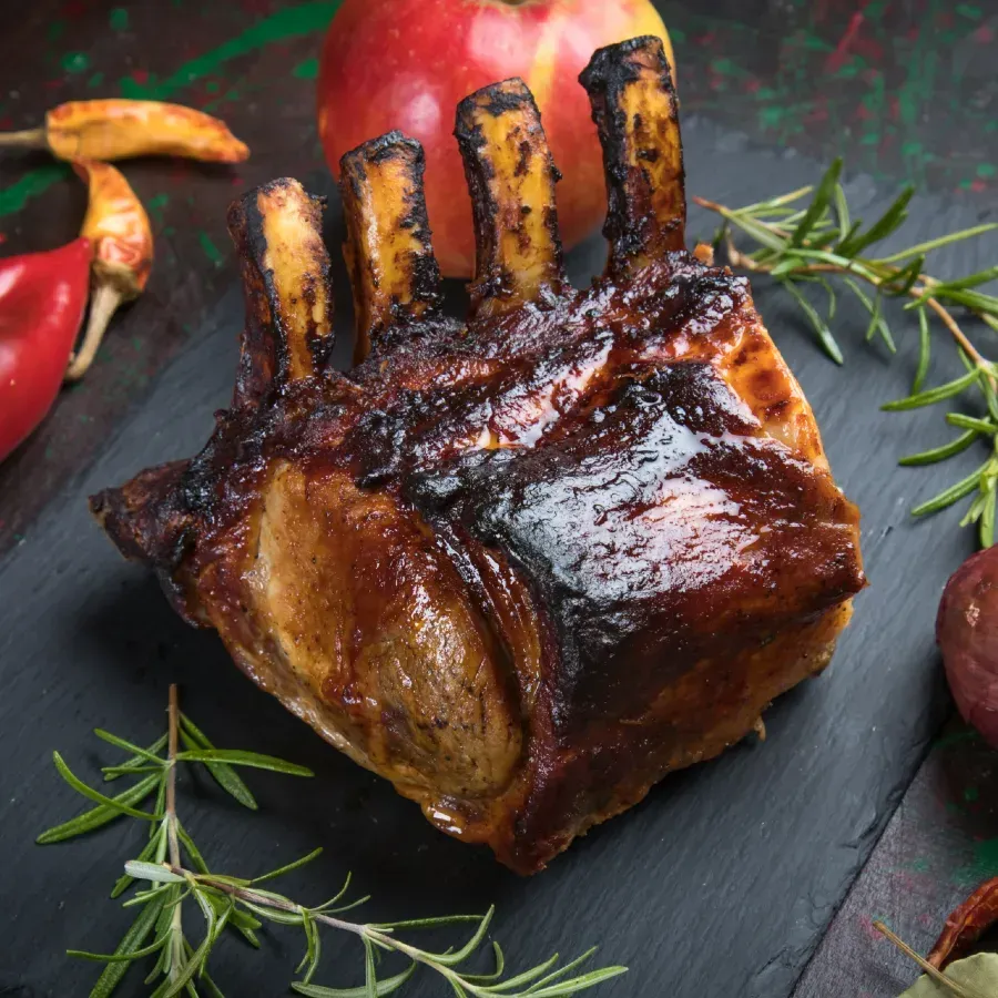Roasted pork rack with visible ribs on a black slate, surrounded by herbs, apple, and peppers.