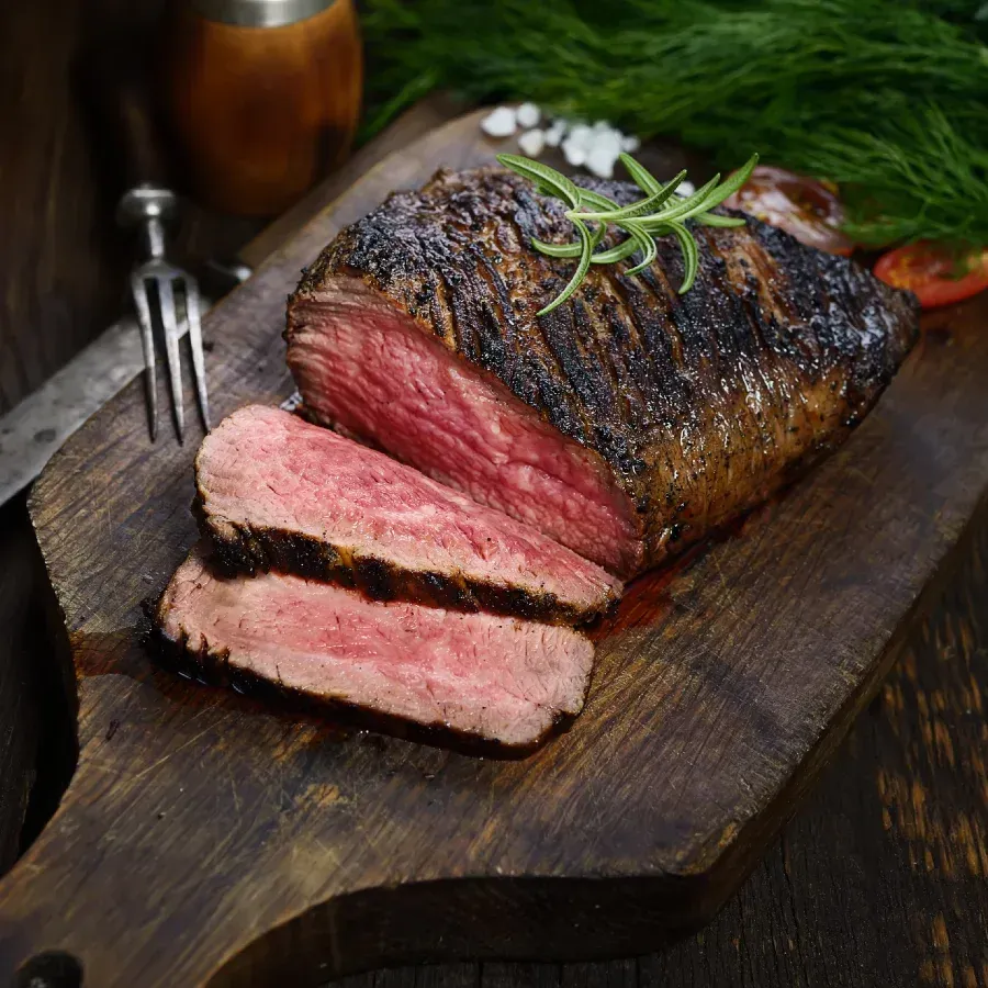 Sliced, medium-rare steak on a wooden cutting board with herbs, salt, knife, and fork.