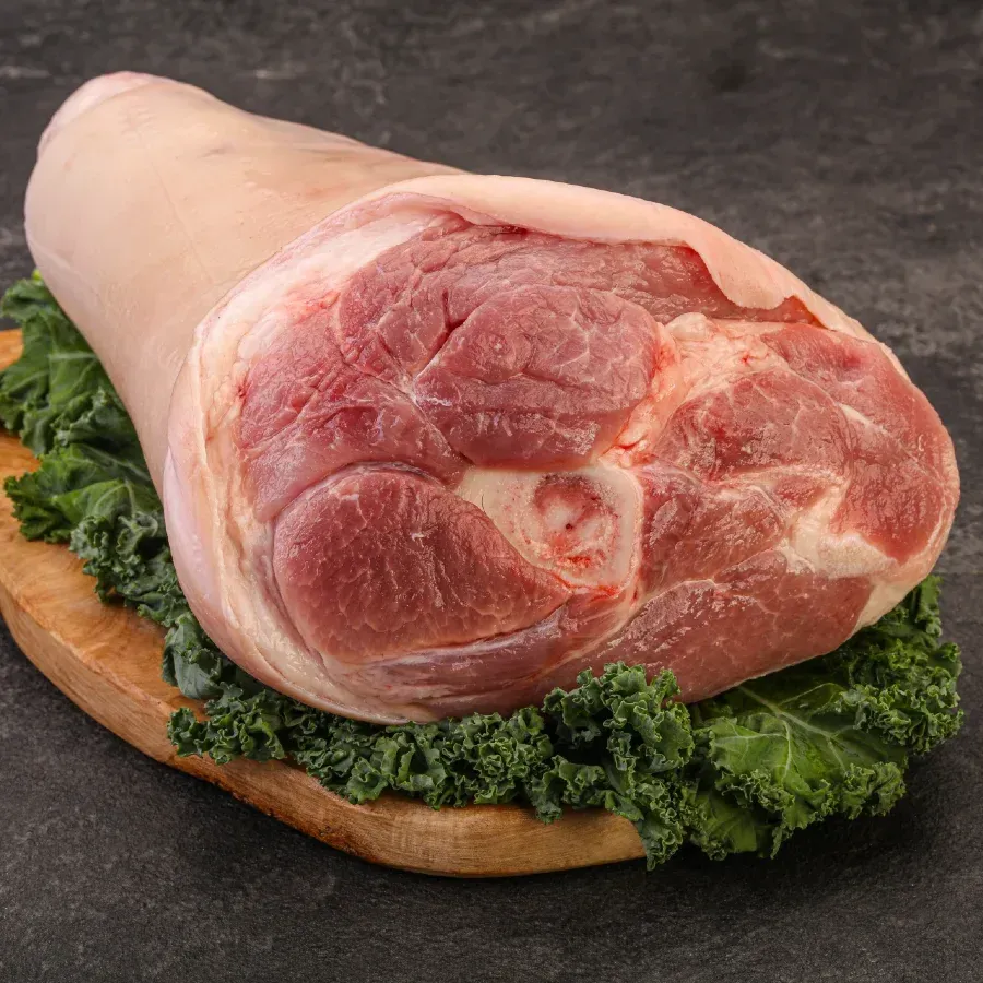 Raw pork knuckle on a wooden board with green kale, against a dark, textured backdrop.