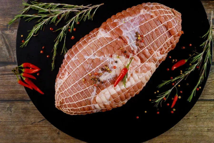 Raw pork roast in netting with herbs and red peppers.