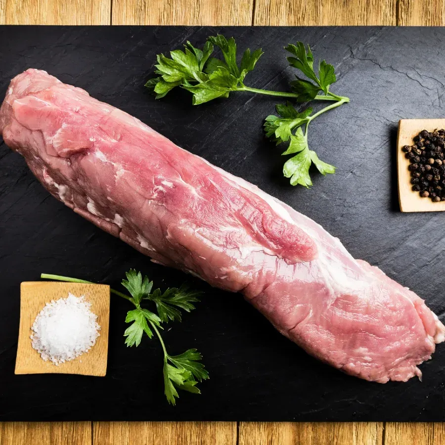 Raw pork tenderloin on a black surface, garnished with parsley, salt, and peppercorns.