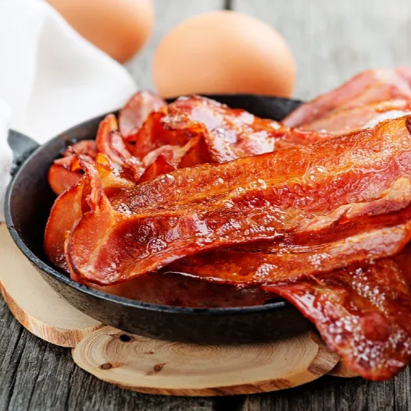 Crispy bacon in a black skillet, two eggs in the background, on a wooden surface.