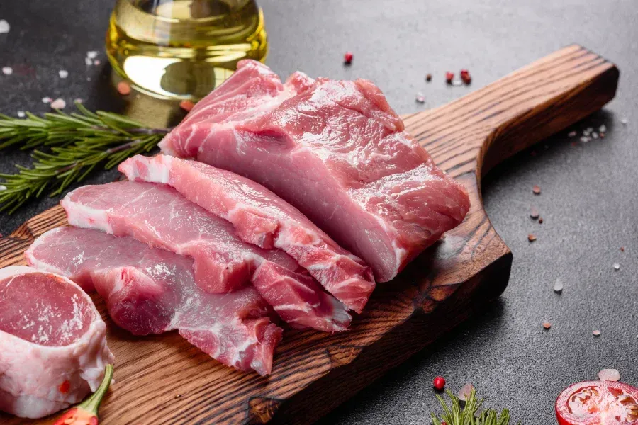 Raw pork loin slices on a wooden cutting board with rosemary, oil, and spices.