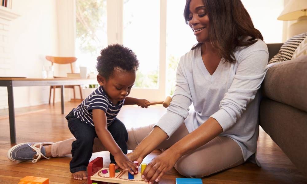 A mother and son are smiling and laughing while playing with developmental blocks and toys on the floor at home.