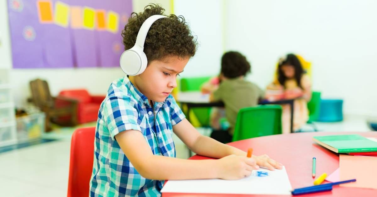 A child sitting at his classroom desk coloring while wearing white headphones.