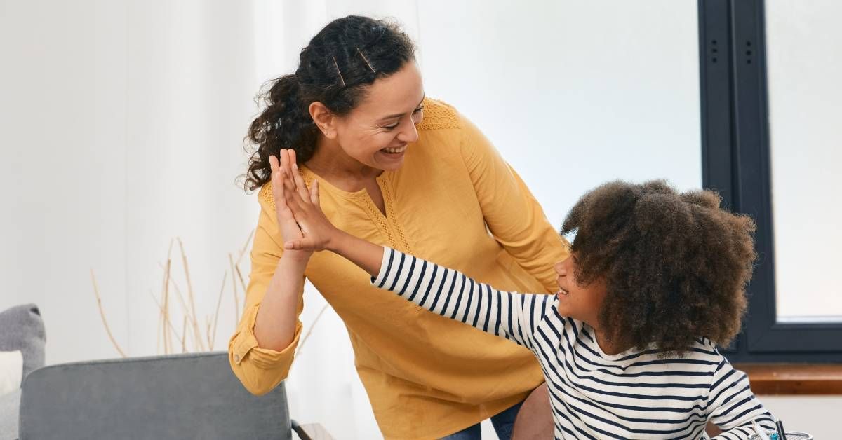 A little girl sitting at a desk with school supplies while giving a woman a high-five. Both are smiling.