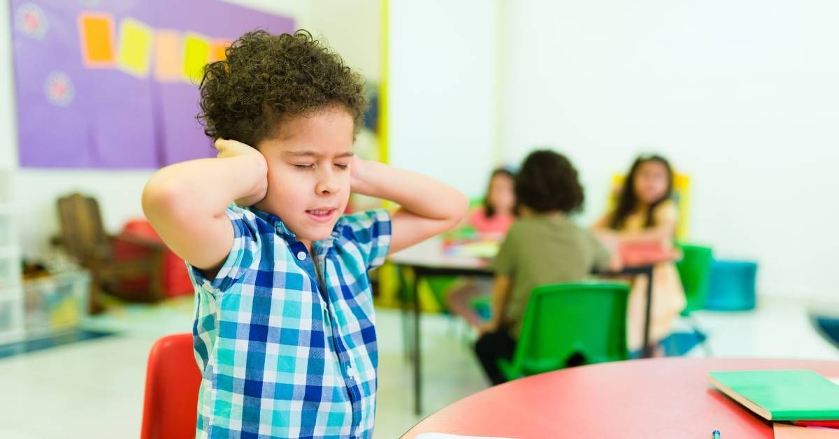 An overwhelmed little boy covering both ears while sitting at a classroom desk.