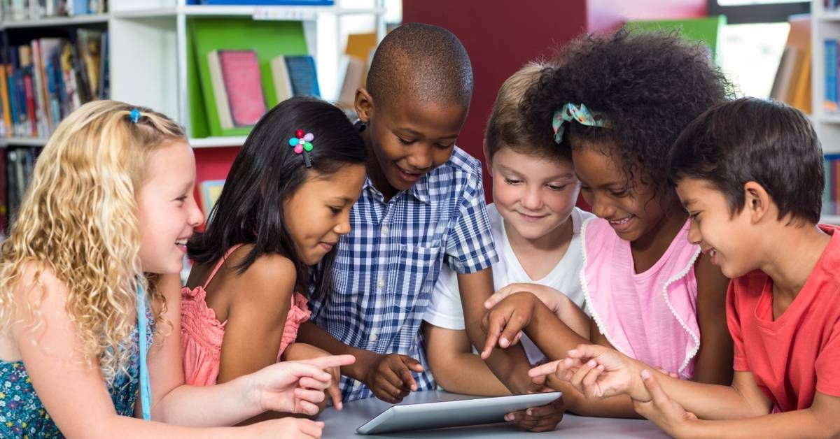 A group of six young children huddled together using a tablet on a desk. They all look amused, and many are smiling.