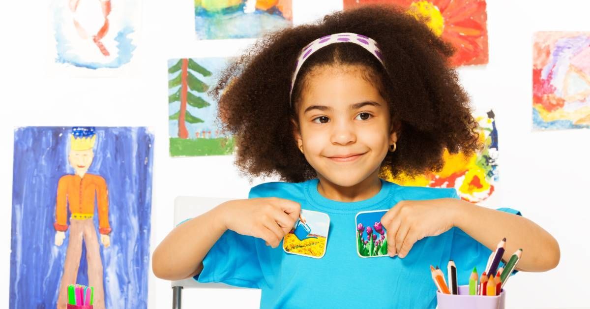 A little girl in blue holding up pictures in each hand while sitting at a desk. There is kid-made art in the background.