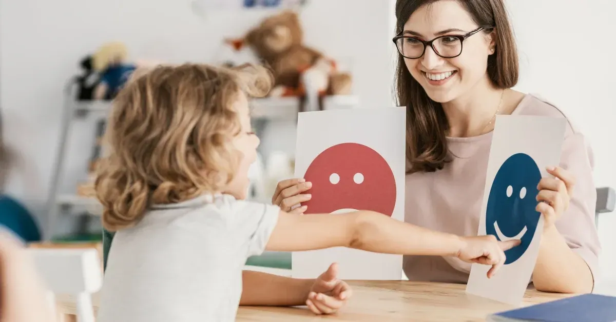 A close-up of a child pointing at a happy emotional emoticon during a therapy session with a psychologist.