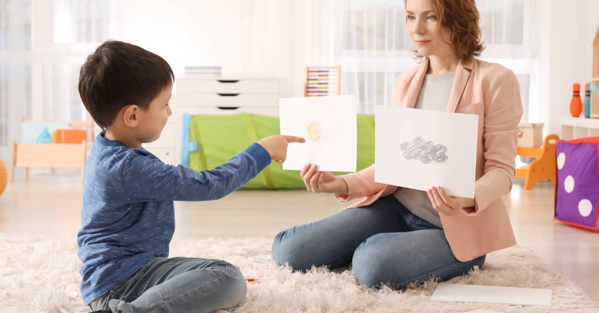 A female psychologist is holding two pictures and working with a boy who is pointing at the cloudy shape.