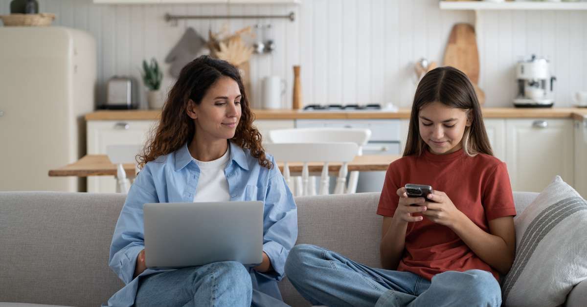 A mom and daughter sitting on a couch in a living room using their electronics.