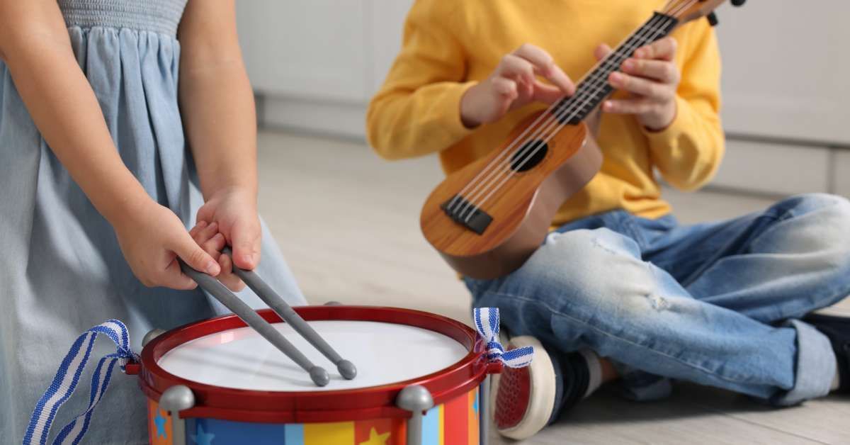 A close-up of two child siblings playing instruments indoors. One is playing the drums, and the other is playing the guitar.