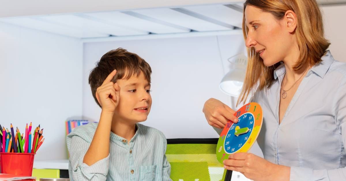 A boy sitting at a desk and learning how to tell time from his teacher. She points at the colorful clock in her hands.