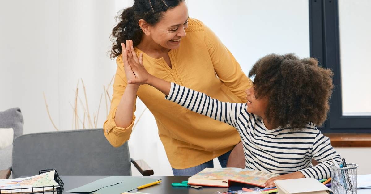 A child and her therapist high-fiving each other and smiling during a tutoring session. Educational materials fill the desk.