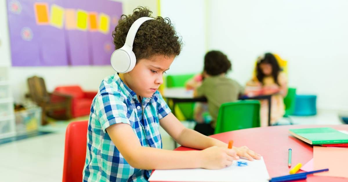 A preschooler with headphones coloring on a piece of paper at school or education center. There are other kids in the back. 