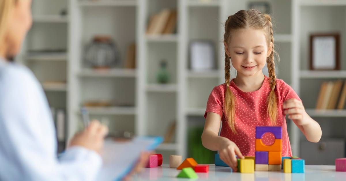 A little girl making a pyramid from colorful wood blocks during a meeting with a teacher or therapist.