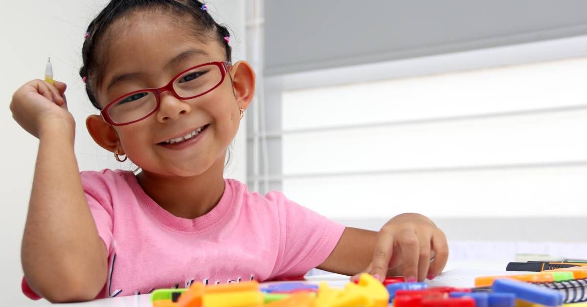 A very young Asian girl smiling with a pencil in her hands while doing homework.