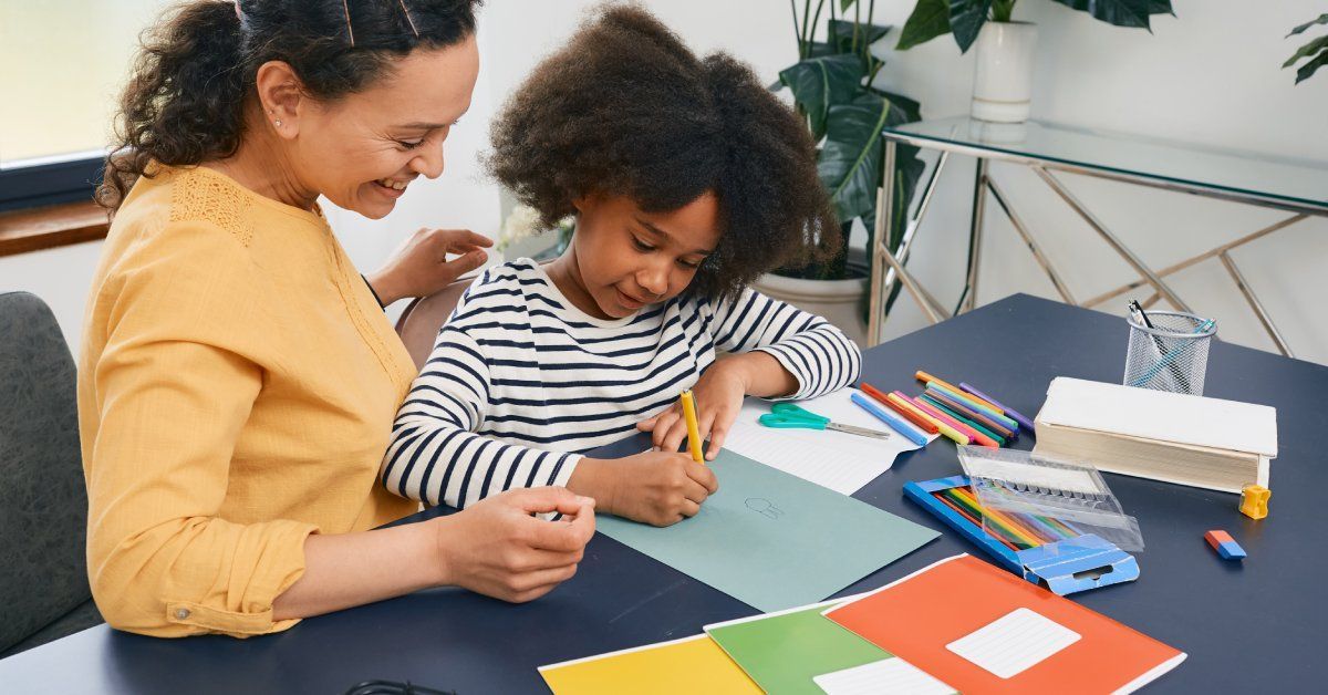 A young girl wearing a striped shirt is sitting with an adult and coloring on a piece of paper. They are both smiling.