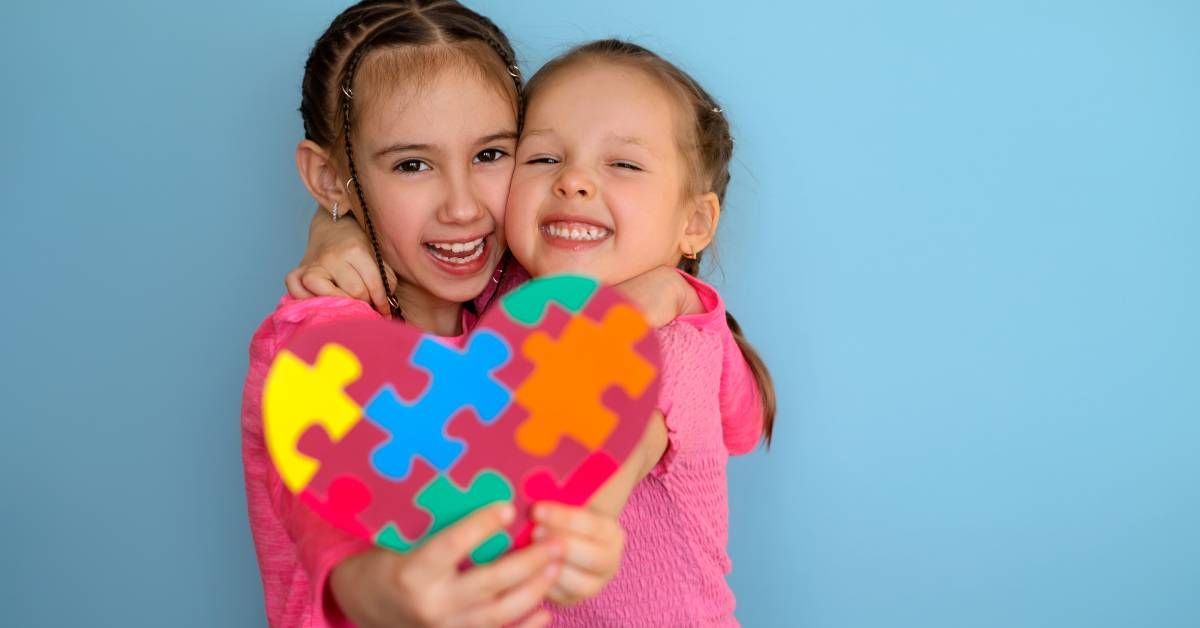 Two young girls hug each other and laugh while holding a heart-shaped paper.