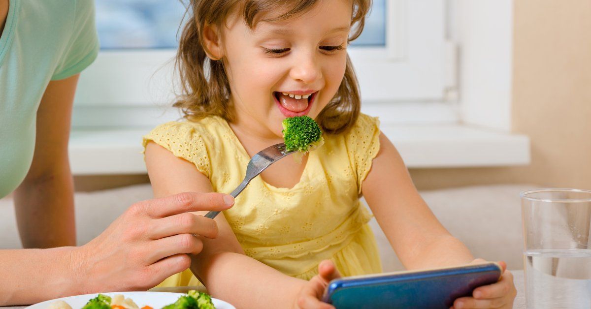 A girl laughs and uses her tablet while her parent feeds her broccoli.