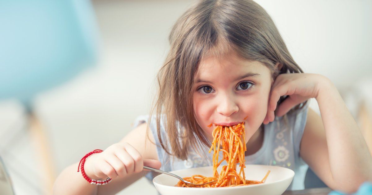 A young girl is sitting at the dinner table and eating pasta with red sauce.