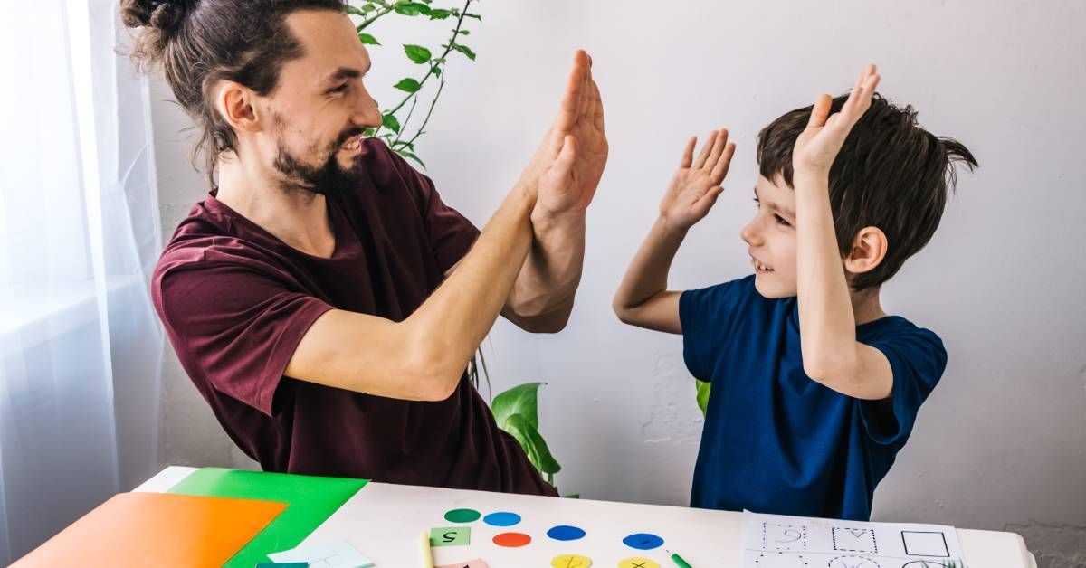 A little boy smiling and high-fiving a man while sitting at a desk. They have art supplies in front of them.