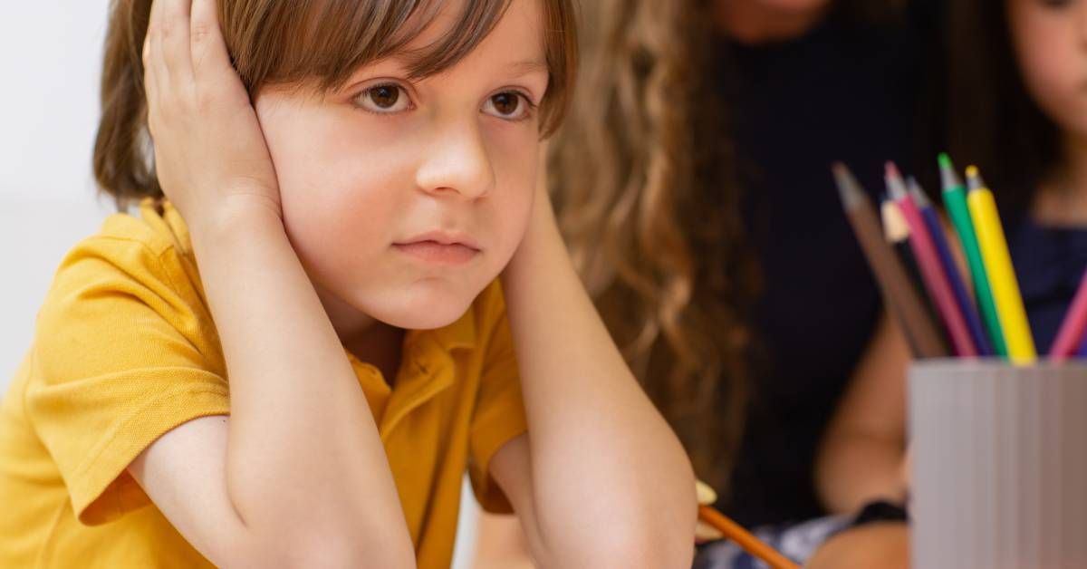 A little boy wearing a yellow shirt is covering both his ears while looking disgruntled. There are art supplies nearby.