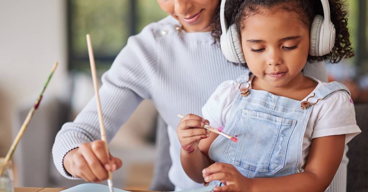 A young girl sits on a woman's lap while the two paint. She wears headphones.