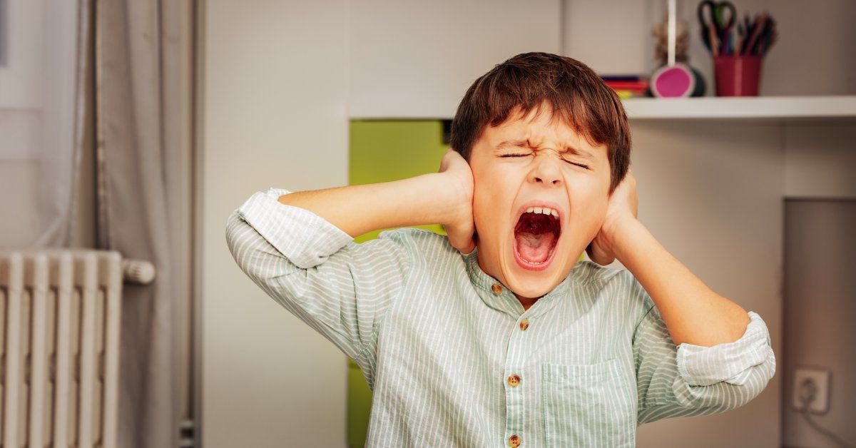 A little boy yelling while squeezing his eyes shut and covering both ears. He sits in a bedroom.