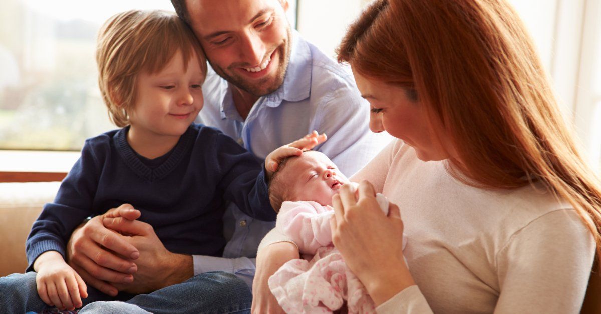 A man holds a young child while a woman holds a newborn baby. All are smiling, and the young boy is touching the newborn's head.