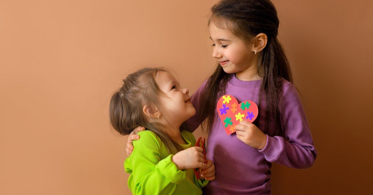 Two young girls hugging while smiling at one another. One holds a paper heart.