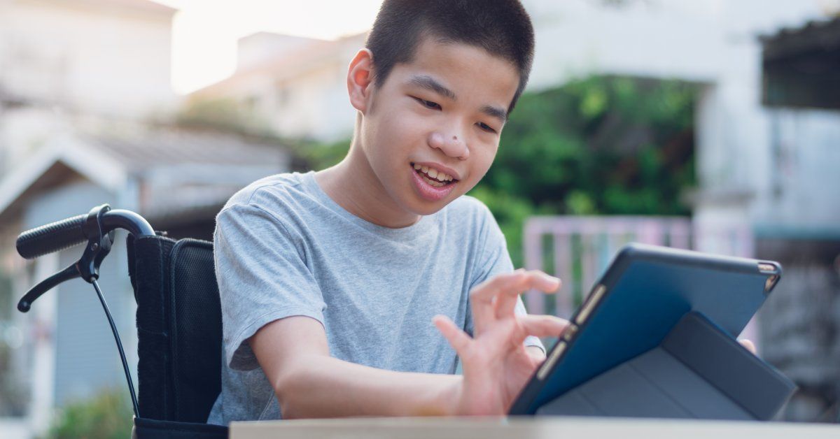 A child in a wheelchair smiling while using a tablet outside on a clear day.