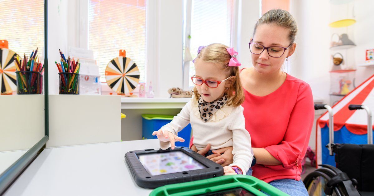 A young girl sits in the lap of a woman while using a tablet screen in a classroom. The girl wears glasses and a leopard-printed scarf.