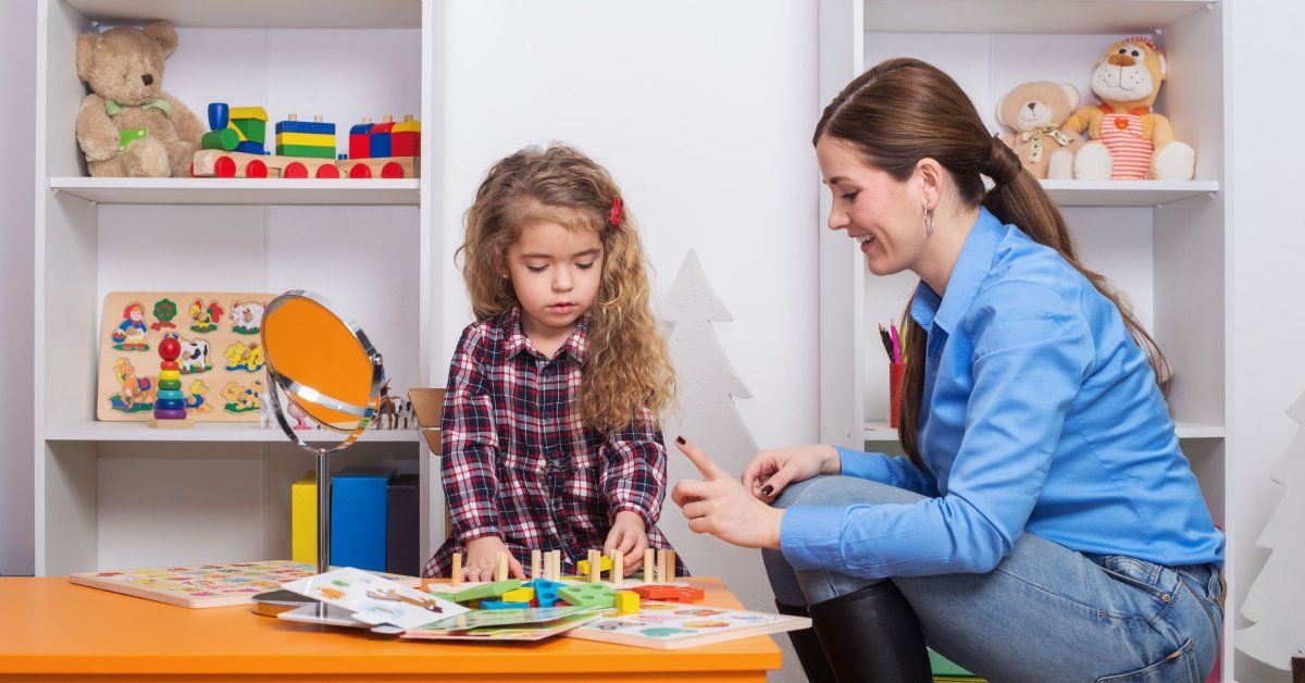 A toddler girl plays with blocks on a desk beside a woman. Shelves with toys are in the background.