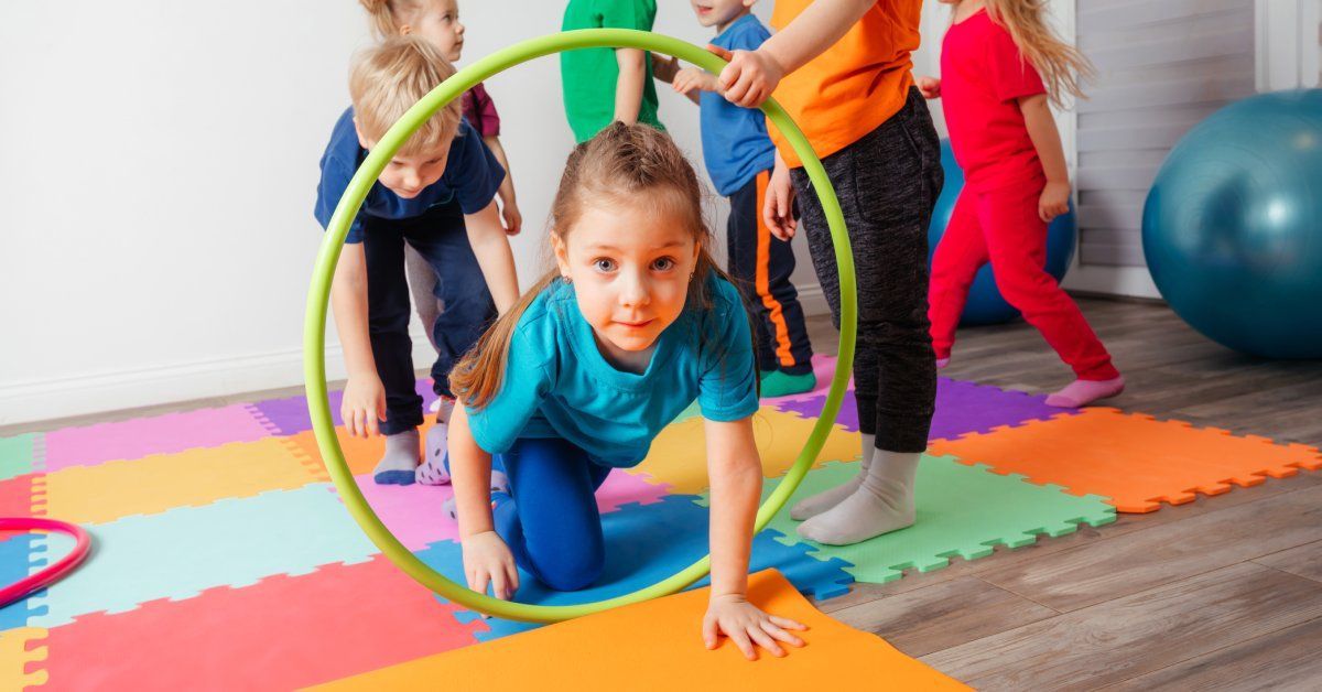 A group of seven young children take turns bending down and walking through a small green hoop.