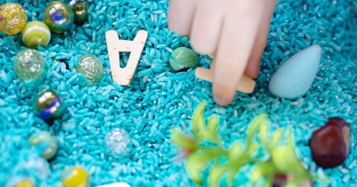 A child's small hand reaches into a sensory bin filled with blue beads, marbles, and letters.